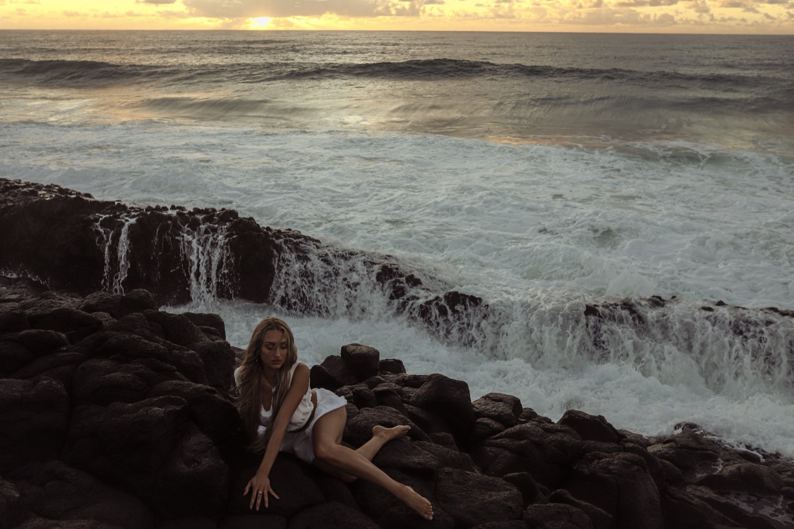 Shanti Griffin seated on volcanic rock beside a tidal waterfall at sunset